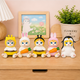 Five cute cartoon animal figurines sitting on a wooden surface in a styled room setting. The figures include a cat in a yellow bee costume, a rabbit in a white bunny suit, a bee in a yellow bee costume, a rabbit in a pink bunny suit holding a carrot, and a penguin with a yellow face mask holding a carrot.