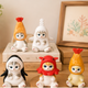 Four small cat figurines dressed in colorful animal costumes, arranged on and around stacked books on a wooden surface in a styled indoor setting with a plant and framed artwork in the background.